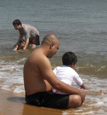 3 photos show an Arab man sitting in the shallow water, and deeper in the water is a Chinese man squatting.  Each man is with a little girl about 2-3 years old.  The Arab girl first stands behind her father, then sits on his lap.  The Chinese father is holding the girl and pulling her through the water.